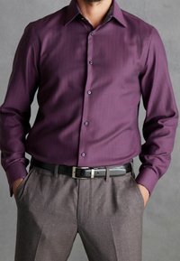 Mann trägt ein eng anliegendes langärmeliges Hemd mit violettem Muster, in graue Hosen mit schwarzem Leder Gürtel gesteckt, Hände in den Taschen.