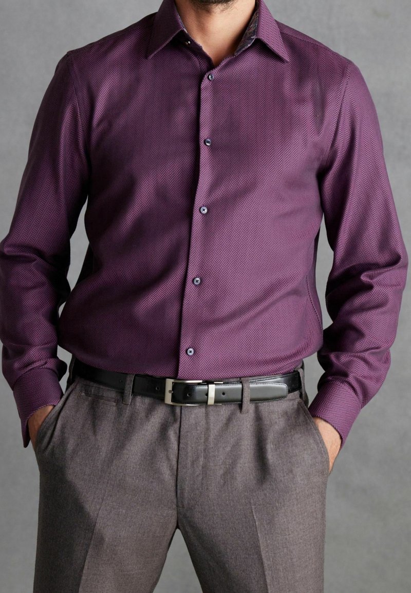 Mann trägt ein eng anliegendes langärmeliges Hemd mit violettem Muster, in graue Hosen mit schwarzem Leder Gürtel gesteckt, Hände in den Taschen.