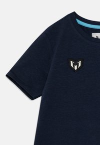 Marineblå t-skjorte med rund hals, korte ermer og en sydd logo-patch med et skjolddesign i svart og hvitt.