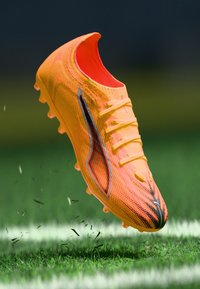 Botín de fútbol de color naranja brillante con un upper texturizado, acentos negros y seis tacos moldeados, suspendido sobre césped verde con pequeños restos.