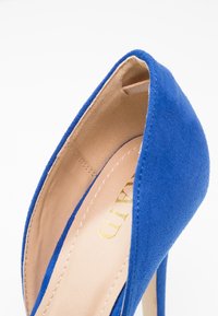 Chaussure à talon haut bleue en daim avec un bout pointu et un intérieur beige lisse, présentant un logo doré subtil et des détails cousus.