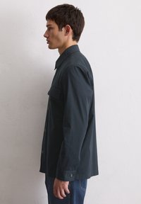 Dunkelgrünes Langarmhemd aus leichtem Stoff, mit durchgehender Knopfleiste, Taschen auf der Brust und einem strukturierten Kragen.
