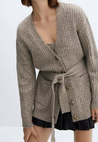 Cardigan côtelé beige avec ceinture à nouer, manches longues et boutons en bois. Comprend deux poches avant. Porté sur une robe noire.