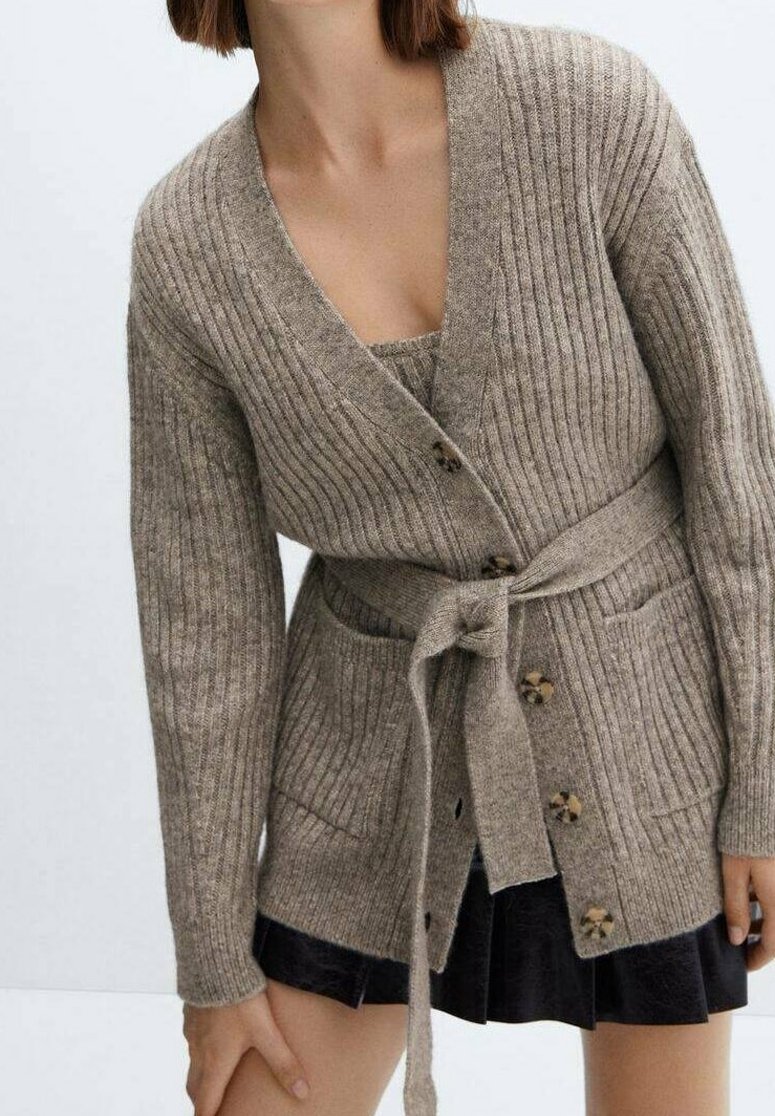 Cardigan côtelé beige avec ceinture à nouer, manches longues et boutons en bois. Comprend deux poches avant. Porté sur une robe noire.