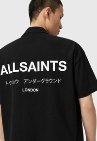 Juodas medvilninis marškinėliai su trumpomis rankovėmis, ant nugaros užrašytas didelis baltas tekstas, įskaitant "ALLSAINTS", japonų ženklus ir "LONDON".
