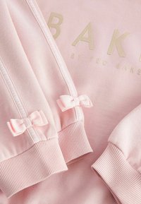 Rosa Sweatshirt mit goldenem "BAKER"-Logo, ausgestattet mit strukturiertem Stoff, gerippten Bündchen und kleinen rosa Schleifenakzenten an den Ärmeln.