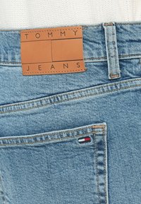Μπλε τζιν παντελόνι με καφέ δερμάτινη ετικέτα που γράφει "TOMMY JEANS" στη μέση. Διαθέτει μια μικρή κόκκινη, λευκή και μπλε λεπτομέρεια σε μια πίσω τσέπη.