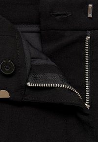 Vêtement noir partiellement dézippé montrant les dents argentées de la fermeture éclair métallique, un bouton noir et une fermeture métallique à crochet et barre sur le tissu.