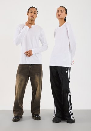 Two people with braided hair wearing white long-sleeve shirts; one in faded brown jeans and boots, the other in black Adidas track pants and shoes.