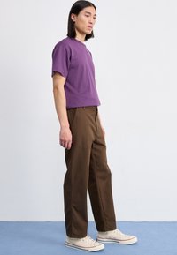 Paarse t-shirt met korte mouwen, gecombineerd met bruine rechte broek en beige sneakers, staande tegen een lichte achtergrond.