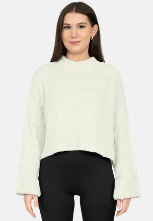 Junge Frau mit langen braunen Haaren, die einen weißen, gerippten Strickpullover und schwarze Hosen trägt, und frontal vor einem schlichten Hintergrund steht.