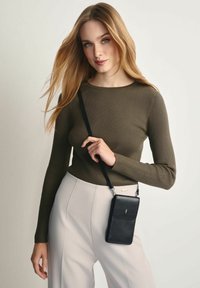 Czarna, teksturowana torebka na ramię o eleganckim, prostokątnym kształcie, noszona na czarnym pasku. Model ma na sobie dopasowaną oliwkowozieloną bluzkę oraz jasne spodnie.