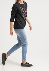 Sudadera negra con diseño floral colorido, combinada con jeans ajustados de mezclilla azul claro y zapatillas negras. Tejido suave, ajuste casual, estilo minimalista.