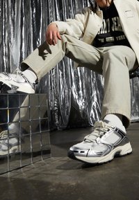 Srebrno-białe sportowe sneakersy z siateczkowymi i gumowymi detalami. Masywna konstrukcja, okrągły czubek i sznurówki. Ustawione na refleksyjnej powierzchni.