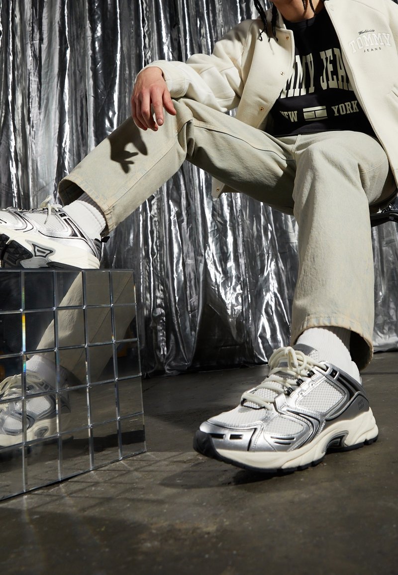 Srebrno-białe sportowe sneakersy z siateczkowymi i gumowymi detalami. Masywna konstrukcja, okrągły czubek i sznurówki. Ustawione na refleksyjnej powierzchni.