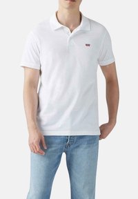 Camisa polo blanca de algodón con cuello, mangas cortas y un pequeño logo rojo de Levi's en el lado izquierdo del pecho. Combinada con unos jeans de color azul claro.