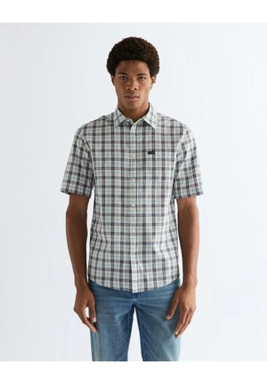 Junger Mann trägt ein kurzärmeliges, kariertes Hemd mit Knöpfen in Blau-, Grün- und Weißtönen, kombiniert mit hellblauen Jeans, stehend vor einem weißen Hintergrund.