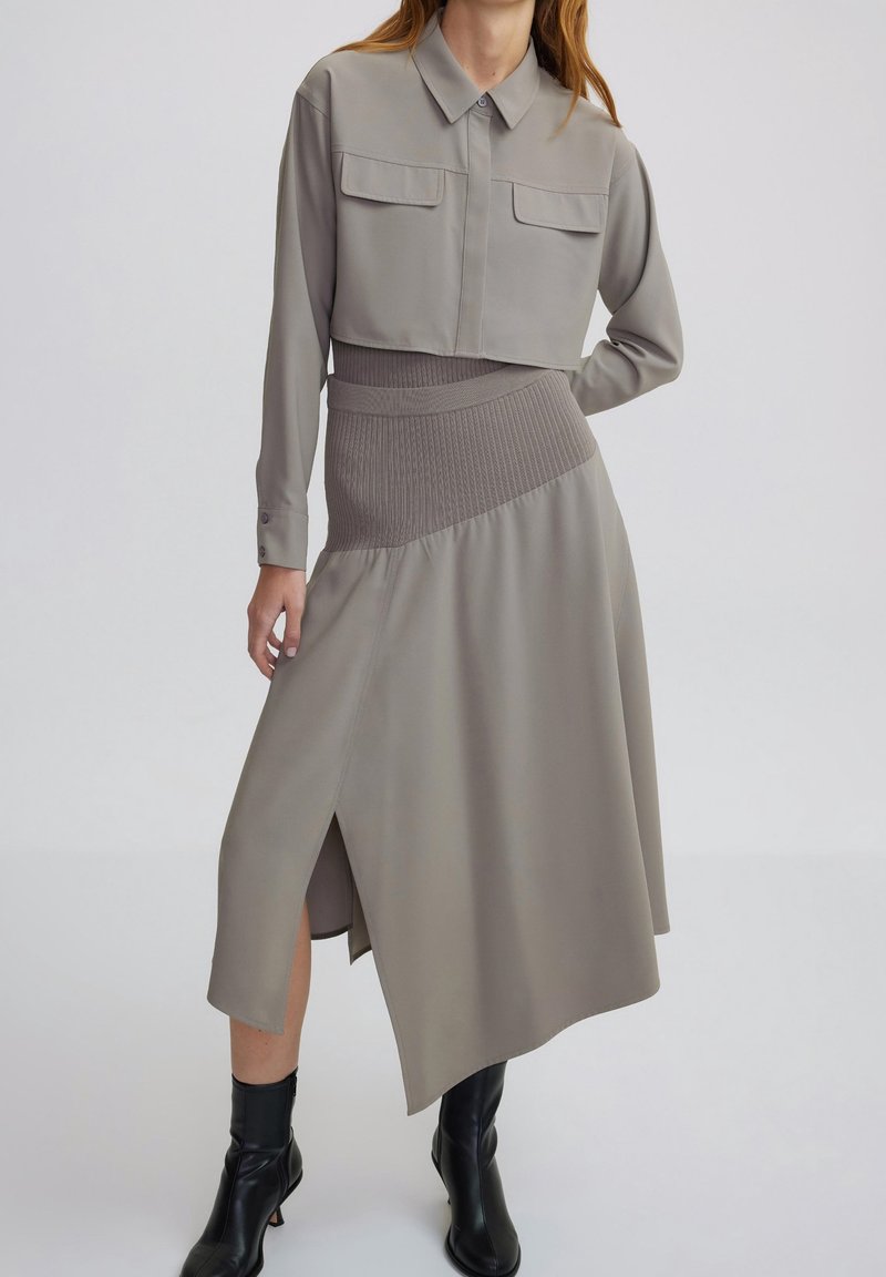 Frau trägt ein taupe-farbenes, kurzes Hemd mit Knöpfen und einen passenden asymmetrischen Midi-Rock mit Schlitz, kombiniert mit schwarzen Knöchelschuhen.
