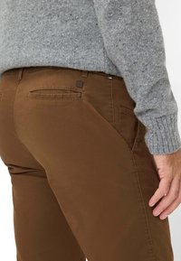 Pantaloni di cotone marroni a gamba dritta, con una tasca posteriore e dettagli di cucitura discreti. Abbinati a un maglione di maglia grigia.