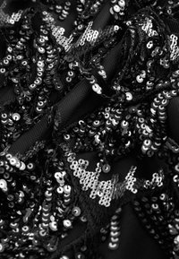 Zwarte stof versierd met dicht op elkaar genaaide zwarte en zilveren pailletten; bevat verschillende vormen en texturen, wat een glinsterend, gelaagd effect creëert.