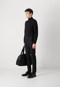 Melnā turtleneck džemperis, pielāgotas melnas bikses un melni vakara apavi, tur melnu sporta somu. Gluda tekstūra un minimālisma dizains.