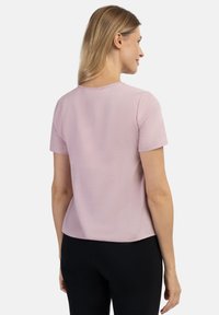 Halenka s krátkým rukávem v měkké růžové barvě, hladká textura, jednoduchý kulatý výstřih a mírně volný střih, nošena s černými kalhotami.