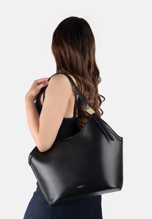 Žena s dugom smeđom kosom drži veliku crnu kožnu torbu preko desnog ramena s zlatnim detaljima, nosi tamni top bez rukava.