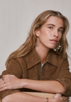 Young woman with long blonde hair wears large gold hoop earrings with charms and a brown suede jacket, resting arms on a surface.