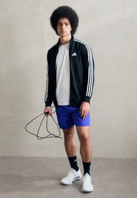 Crna sportska jakna s bijelim prugama, bijela majica, plave kratke hlače, bijeli tenisice, drži crni konopac za preskakanje na neutralnoj pozadini.