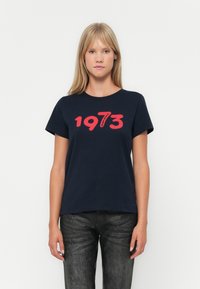 Tumši zila kokvilnas T-krekls ar lielu sarkanu uzdruku "1973" uz krūšu. Īsas piedurknes un apaļa kakla izgriezuma forma.