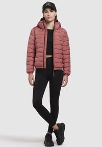 Padded pink jacket with hood, quilted design, front zipper, paired with black leggings and black sneakers; model stands with arms relaxed.