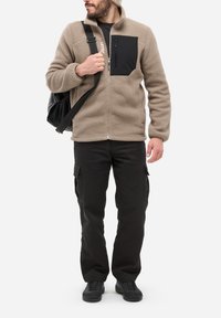 Homme portant une veste en polaire beige avec une poche poitrine noire, un pantalon cargo noir, des chaussures noires, un bonnet en tricot beige, et portant un sac à bandoulière noir.