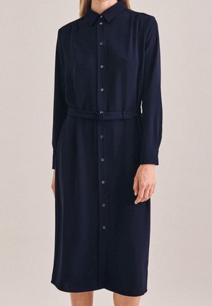 Femme portant une robe midi bleu marine à manches longues, boutonnée, avec un col et une ceinture assortie à la taille.