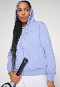 Kvinne i en lyse lilla zip-up hettegenser som holder en tennisracket, og ser til siden med lange flettede hår over skulderen.