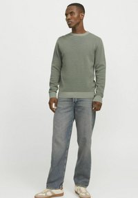 Grüner strukturierter Pullover mit gerippten Bündchen und Saum, getragen mit hellblauen Jeans und beigen Turnschuhen, vor einem neutralen Hintergrund stehend.