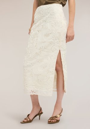 Mujer con falda midi blanca texturizada con una abertura lateral y sandalias marrones de tacón con tiras, de pie sobre un fondo liso.