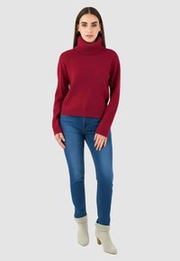 Maglione a collo alto color bordeaux con polsini e orlo a costine, abbinato a jeans blu slim fit e stivaletti beige chiaro alla caviglia.