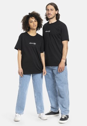 Zwei Personen tragen passende schwarze Baumwoll-T-Shirts mit weißem Text, kombiniert mit hellblauen Jeans und kontrastierenden Turnschuhen.