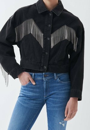 Person trägt eine schwarze kurz geschnittene Jacke mit silbernen Fransen über der Brust und an den Ärmeln, kombiniert mit blauen, abgenutzten Jeans.