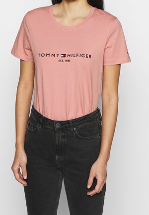 Femme portant un t-shirt rose Tommy Hilfiger avec logo et texte "EST. 1985", associé à un jean noir taille haute, debout devant un fond blanc.