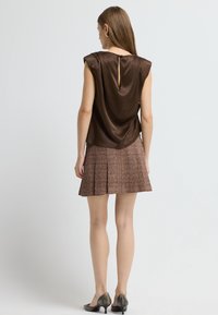 Brun satin uden ærmer med en nøglehul-bagdetalje, parret med en plisseret brun tweednederdel. Sættet har subtile teksturer og farver.