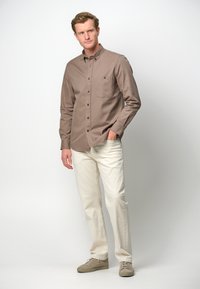Bruine overhemd met knoppen en een borstzak, gecombineerd met lichtgekleurde broek en beige sneakers, tegen een witte achtergrond.