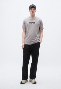 Γκρι κοντομάνικο μπλουζάκι με μαύρο λογότυπο "BOSS", συνδυασμένο με μαύρο παντελόνι και ανοιχτόχρωμα αθλητικά παπούτσια. Το μοντέλο στέκεται μπροστά σε λευκό φόντο.