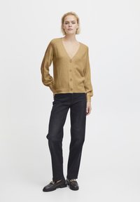 Beige Strickjacke mit V-Ausschnitt, fünf Knöpfen vorne und langen Ärmeln. Kombiniert mit schwarzen geraden Hosen und schwarzen Schuhen.