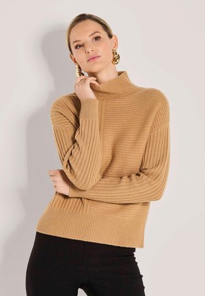 Frau in einem beigen, gerippten Rollkragenpullover und schwarzen Hosen, mit goldenen auffälligen Ohrringen, posiert mit einer Hand, die ihr Gesicht berührt.