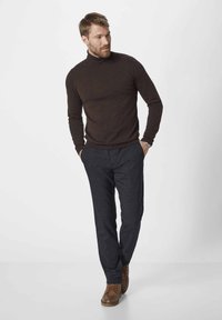 Brown turtleneck sweater, textured. Dark patterned trousers, loose fit. Tan leather shoes with laces. Model stands with hands in pockets.