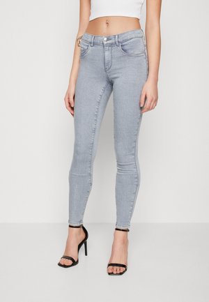Kvinde iført lysegrå højtaljede skinny jeans og sorte åbne højhælede sandaler, stående foran en ensartet hvid baggrund.