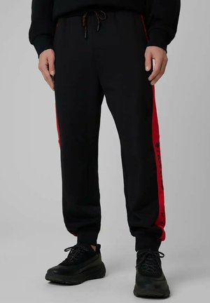 Mężczyzna w czarnych dresach z czerwonymi paskami po bokach i czarnych trampkach, stojący na jednolitym szarym tle.