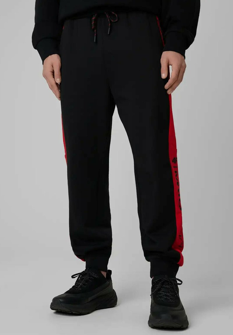 Homme portant un pantalon de survêtement noir avec des bandes latérales rouges et des baskets noires, se tenant devant un fond gris uni.