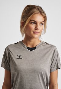 Grå atletisk t-shirt med korte ærmer, med en v-hals og sort logo på brystet. Blød, tekstureret stof med en let glans.